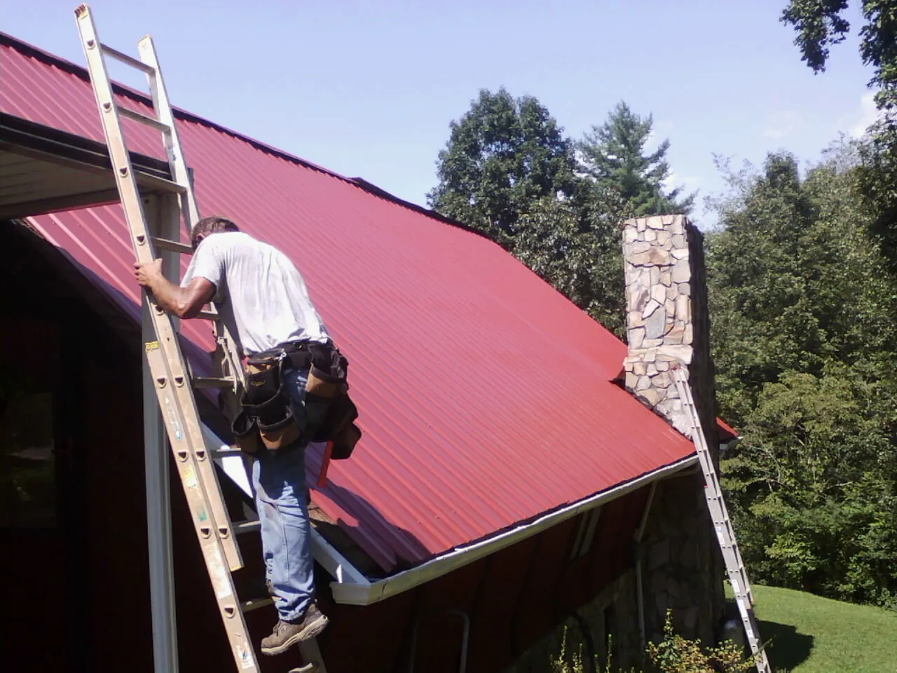 Roofer on a ladder beside a red metal roof and stone chimney — Soffit & Fascia Repair by Roofing Brothers in Rockville